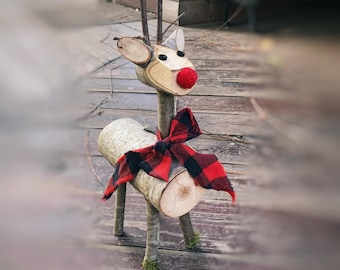 May include: A wooden reindeer figurine with a red pom-pom nose and a red and black plaid bow. The reindeer is standing on a wooden surface.