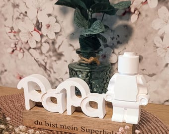 May include: A white wooden "Papa" sign with a matching figurine, set on a wooden base with the text "du bist mein Superheld". A green glass vase holds greenery. The background features a floral wallpaper.