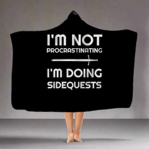 May include: Black hooded blanket with the text "I'm not procrastinating I'm doing sidequests" in white lettering with a sword graphic.