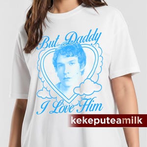 May include: White t-shirt with a blue graphic. The graphic features a portrait inside a heart-shaped frame with the text "But Daddy I Love Him". The brand name "kekepteamilk" is printed at the bottom.