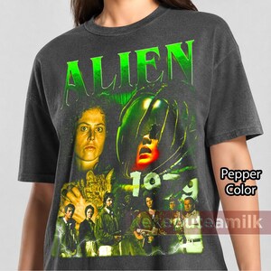 May include: A charcoal gray t-shirt featuring a graphic print from the movie Alien. The design includes the word ALIEN in bright green letters, a portrait of a character, a cat, and a group of people. The text 1979 is visible in green.