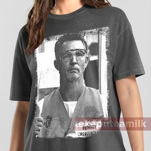 May include: Dark gray t-shirt featuring a black and white image of a man wearing glasses and a vest. The image is framed with a white border. The text "PRIMARY EMERGENCY" is visible on the shirt.