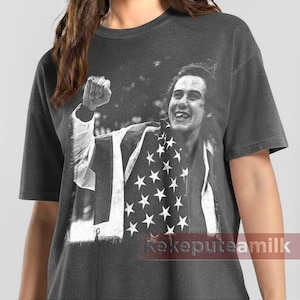 May include: Dark gray t-shirt featuring a black and white image of a person holding an American flag. The person is smiling and has their fist raised in the air. The shirt has a vintage, distressed look.