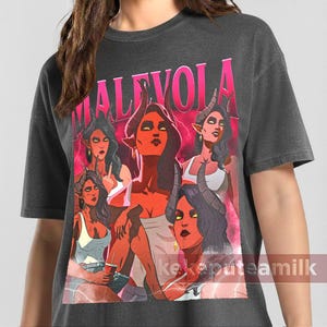 Puede incluir: Camiseta gris oscuro con un gráfico de varias figuras femeninas con cuernos y piel roja, con la palabra "MALEVOLA" en rosa. El diseño tiene un aspecto vintage y desgastado.