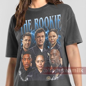 May include: Dark grey crew neck t-shirt featuring a group of people with the text "THE ROOKIE" in blue. The image includes a lightning bolt design. The shirt is a crew neck style.