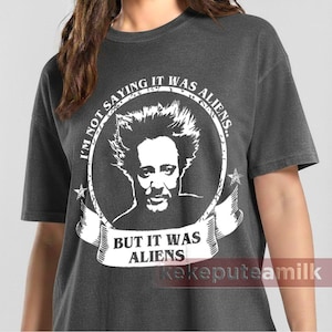 May include: Dark gray t-shirt with a white graphic. The graphic features a portrait inside a circle with the text "I'M NOT SAYING IT WAS ALIENS" and "BUT IT WAS ALIENS". The shirt has short sleeves and a crew neck.