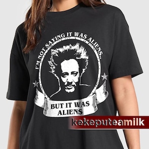 May include: Black t-shirt with a white graphic of a man's face inside a circular frame. The text "I'M NOT SAYING IT WAS ALIENS" arcs around the top, and "BUT IT WAS ALIENS" is on a banner below the face. The brand name "kekeputeamilk" is at the bottom.