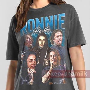 May include: Dark gray t-shirt featuring a graphic of the band Ronnie Radke. The design includes the band's name in blue, with a lightning bolt effect, and images of the band members. The shirt has a vintage, distressed look.