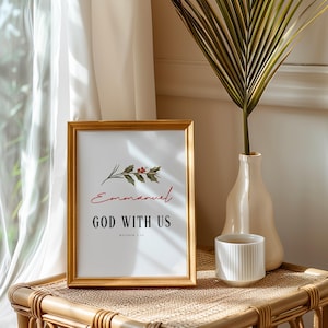 Pode incluir: Uma impressão emoldurada com moldura dourada exibe as palavras "Emmanuel GOD WITH US" em letras pretas e vermelhas. Um raminho de folhas e bagas está acima da palavra "Emmanuel". Um vaso branco com uma folha de palmeira e uma caneca branca estão em uma mesa tecida.