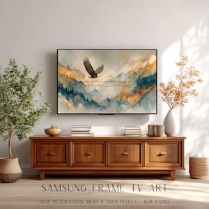 May include: A Samsung Frame TV displaying a watercolor landscape with an eagle soaring over mountains. The artwork includes text from Psalm 103:5. The TV is mounted above a wooden cabinet with drawers, complemented by decorative plants and a vase.