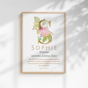 May include: A framed print with the name "Sophie" in gold, decorated with flowers and leaves. The design includes the words "Wisdom, Artistic, Loving, Kind" and a Bible verse. The print is in a wooden frame, suitable for home decor.