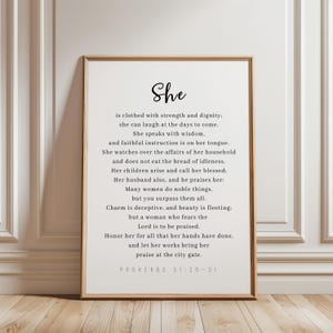 May include: A framed print with the word "She" in elegant script at the top. The print features a passage from Proverbs 31:25-31, written in a classic font, set against a white background. The frame is a warm, light brown.