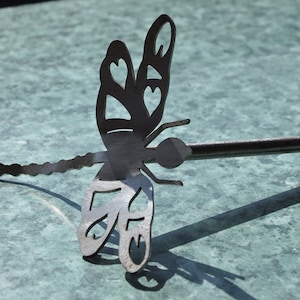 Pode incluir: Uma estaca de jardim de metal em forma de libélula com acabamento preto e desenhos de coração recortados. A estaca é feita de metal e tem um caule longo e fino.