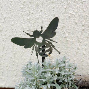 Pode incluir: Uma estaca de jardim de metal em forma de abelha com um coração recortado em seu corpo. A abelha está em um vaso de plantas de folhas prateadas.