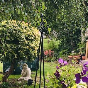 Pode incluir: Um obeliscos de jardim de metal preto com uma parte superior em espiral decorativa. O obelisco está em um jardim com grama verde, uma pequena estátua e várias plantas.