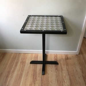 May include: A black metal table with a square top made of diamond plate metal. The table has a single, black metal leg that forms a cross at the base.