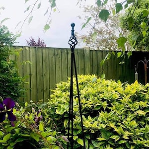 Pode incluir: Um obeliscos de jardim de metal preto com um remate decorativo. O obelisco está situado num jardim com folhagem verde e amarela.
