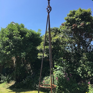 Puede incluir: Un obeliscos de jard&iacute;n de metal oxidado con una parte superior decorativa y un anillo en el medio. El obelisco est&aacute; de pie en un &aacute;rea cubierta de hierba con follaje verde en el fondo.