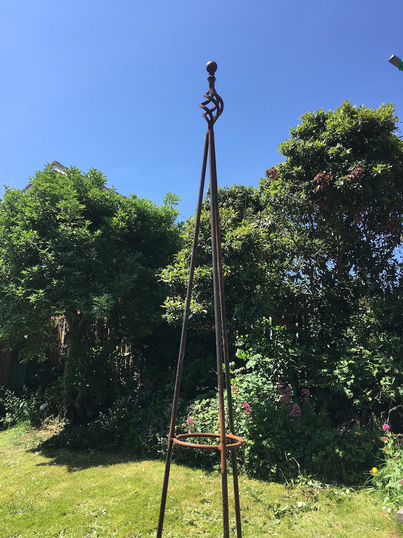 Puede incluir: Un obeliscos de jard&iacute;n de metal oxidado con una parte superior decorativa y un anillo en el medio. El obelisco est&aacute; de pie en un &aacute;rea cubierta de hierba con follaje verde en el fondo.