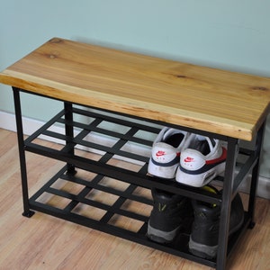 May include: A wooden bench with a black metal shoe rack underneath. The bench has a natural wood finish and the rack has two shelves. There are shoes on the shelves, including a pair of white sneakers with red accents.