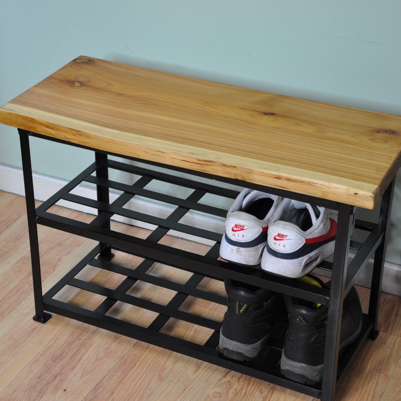 May include: A wooden bench with a black metal shoe rack underneath. The bench has a natural wood finish and the rack has two shelves. There are shoes on the shelves, including a pair of white sneakers with red accents.