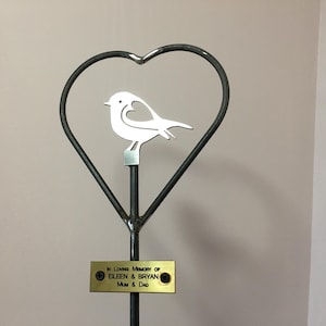 Pode incluir: Uma estaca de jardim de metal em forma de coração com uma silhueta de pássaro de metal branco no topo. A estaca tem uma placa dourada que diz "In Loving Memory of Eileen & Bryan Mum & Dad".