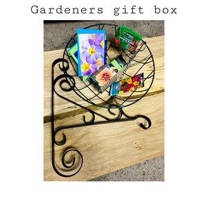 Könnte beinhalten: Ein grüner, hängender Metallkorb gefüllt mit Gartenartikeln, darunter Saatgutpakete, Gartenhandschuhe und eine kleine Karte. Der Korb ist an einer schwarzen Metallhalterung befestigt.