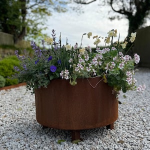 Puede incluir: Una jardinera redonda de metal oxidado llena de una variedad de flores coloridas. Las flores incluyen flores moradas, blancas y amarillas. La jardinera se asienta sobre tres patas cortas y se coloca sobre un lecho de pequeñas piedras grises.