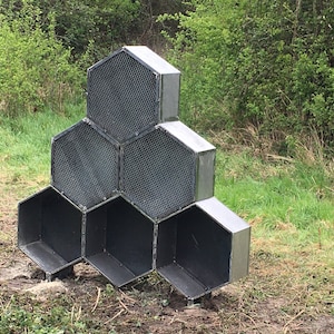 Può includere: Una scultura in metallo a forma di alveare con sei scomparti esagonali. La scultura è realizzata in metallo nero e ha un aspetto grezzo e industriale. È posizionata su un prato erboso.