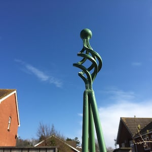 Pode incluir: Um treliça de jardim de metal verde com um topo decorativo. O topo é um design em espiral com um remate arredondado.
