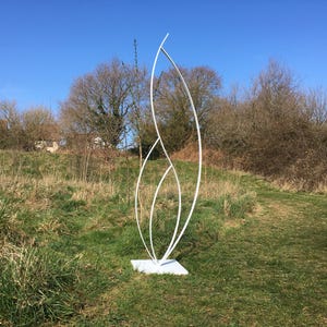 Pode incluir: Escultura de metal branco com design abstrato e fluido. A obra está sobre uma base quadrada num campo gramado, com árvores e céu azul ao fundo. Design moderno e minimalista, ideal para ambientes externos.