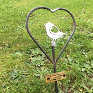 Pode incluir: Um memorial de metal em forma de coração com um recorte de pássaro e uma pequena placa que diz "In Loving Memory of ELIN & BROWN May 8, 1944". O memorial está colocado em uma área gramada.