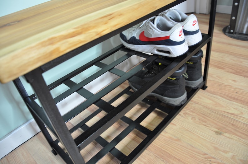 May include: A black metal shoe rack with a wooden top. The rack has three shelves and is filled with shoes. The shoes are a pair of white sneakers and a pair of black work boots.