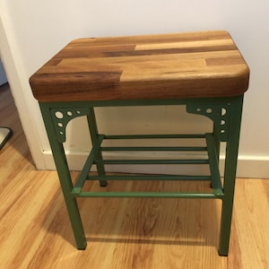 Puede incluir: Taburete con tapa rectangular de madera. El asiento está hecho de tablas de madera marrón clara y oscura. Las patas y el marco están pintados de color verde apagado. El taburete está diseñado para sentarse.