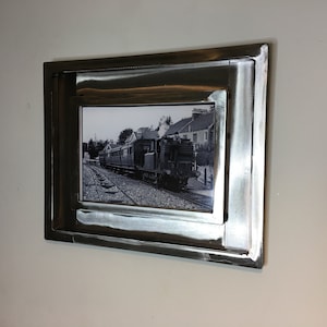 May include: A black and white photograph of a train in a metal frame. The frame has a brushed metal finish and is mounted on a white wall. The photograph depicts a vintage train on tracks.