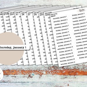 May include: A stack of white date stickers for 2026, with black text. The stickers are arranged on a distressed, light-colored wooden surface. The text includes the months, days of the week, and dates. A tan circle with the text "thursday, january 1" is in the center.