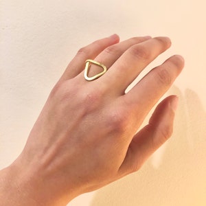 Può includere: Un anello d'oro a forma di triangolo con un centro vuoto. L'anello è indossato sul dito medio di una mano.