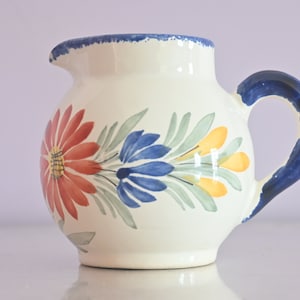 Può includere: Un'anfora di ceramica bianca con manico blu e motivo floreale. I fiori sono rossi, blu, gialli e verdi.
