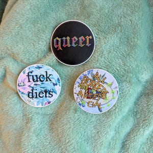May include: Three round stickers with text and designs. One black sticker says "queer" in colorful letters. Another says "fuck diets" with a floral background. The third says "All borders are artificial".