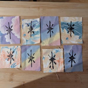 May include: Eight small fabric squares with a black starburst design. The squares have a variety of pastel floral and tie-dye patterns in shades of pink, blue, yellow, and peach. The squares are arranged on a wooden cutting board.