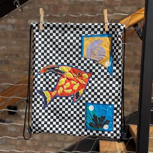 Puede incluir: Mochila con cordón con un patrón de cuadros blancos y negros. La bolsa presenta una aplicación de pez naranja, amarillo y negro. Los parches adicionales incluyen un cuadrado amarillo con un remolino púrpura y un cuadrado azul con un diseño de coral.