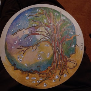 May include: A circular wooden board with a painted design of a tree with roots, branches, and leaves. The tree is surrounded by a blue, purple, and yellow sky with white dots. The board has the text "KL 23" written on the right side.