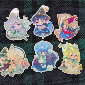 Puede incluir: Colección de seis pegatinas holográficas con personajes de estilo anime en varias poses. Los personajes están representados con colores vibrantes, vistiendo sombreros y túnicas. Las pegatinas tienen un acabado brillante e iridiscente.