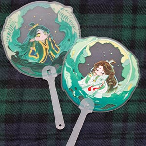 May include: Two round, translucent hand fans with cartoon illustrations. One features a figure in green robes within a wave design, the other shows a figure in white robes. Both have clear plastic handles. The background is a dark plaid.