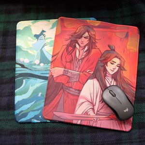 Puede incluir: Dos alfombrillas de ratón con diseños diferentes. La primera alfombrilla de ratón tiene un diseño azul y verde con una figura blanca en el centro. La segunda alfombrilla de ratón tiene un diseño rojo y naranja con dos figuras en el centro. Un ratón de ordenador negro está encima de la segunda alfombrilla de ratón.