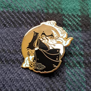 May include: A gold-colored enamel pin featuring a stylized illustration of two figures in traditional clothing, set against a golden moon and white clouds. The figures are primarily black and white, with accents of light blue.