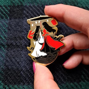 Könnte beinhalten: Eine goldfarbene Emaille-Anstecknadel mit zwei Figuren in einem stilisierten Design. Eine Figur trägt ein weißes Kleid, die andere einen roten Umhang. Die Anstecknadel hat schwarze und rote Akzente mit einem goldenen Rand.