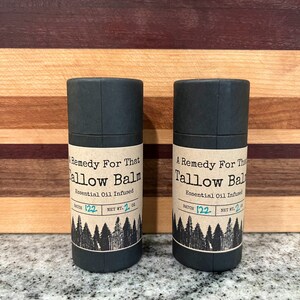 May include: Two dark gray cylindrical containers of Tallow Balm, each with a label featuring text and a forest graphic. The label reads "A Remedy For That Tallow Balm, Essential Oil Infused, Net Wt. 2 oz."