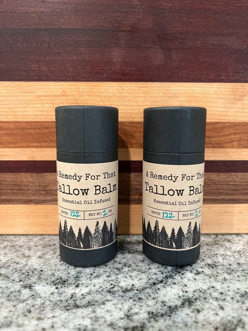 May include: Two dark gray cylindrical containers of Tallow Balm, each with a label featuring text and a forest graphic. The label reads "A Remedy For That Tallow Balm, Essential Oil Infused, Net Wt. 2 oz."