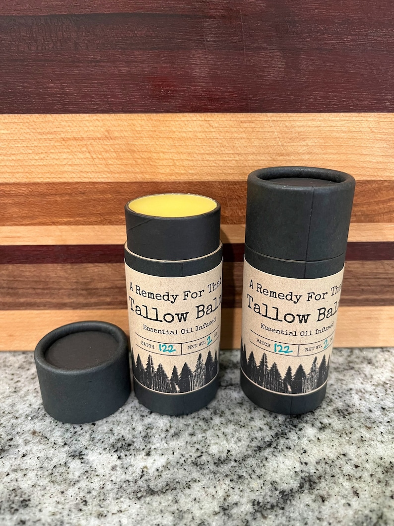 May include: Two black cylindrical containers of tallow balm with the text "A Remedy For This Tallow Balm" and "Essential Oil Infused". One container is open, revealing a yellow balm. The net weight is 2 oz.